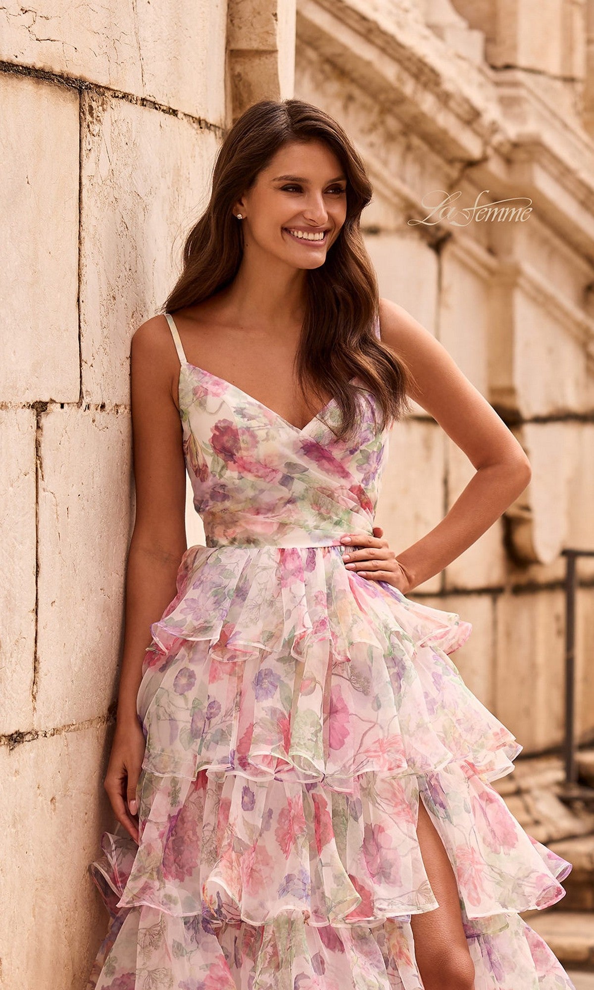 A woman poses smiling against a stone wall in the La Femme 33740 long prom dress—a sleeveless floral chiffon gown with ruffles and a high slit.